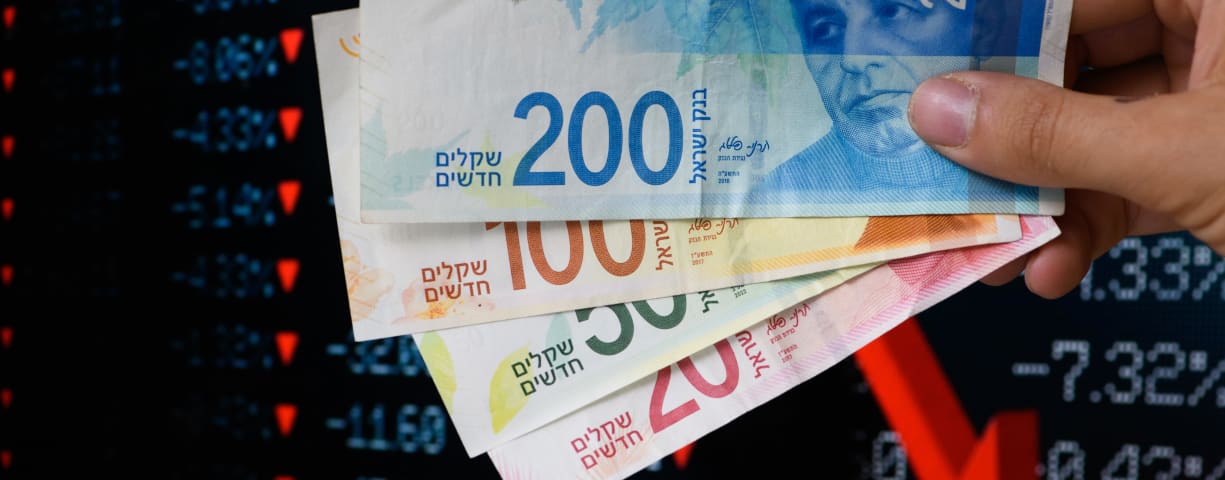 New Israeli Shekel bills are seen in front of a downwards-trending graph (illustration)