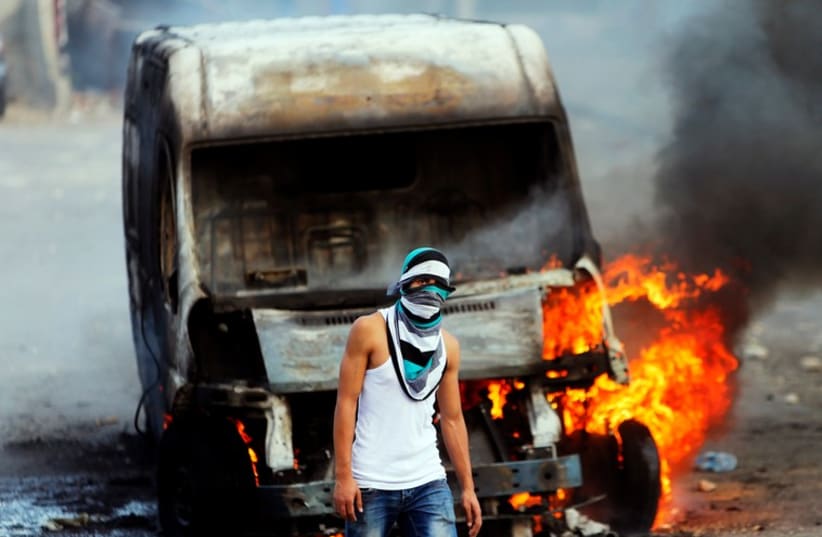 A Palestinian protester stands in front of an Israeli car set torched during clashes with Israeli security forces in east Jerusalem (photo credit: REUTERS)