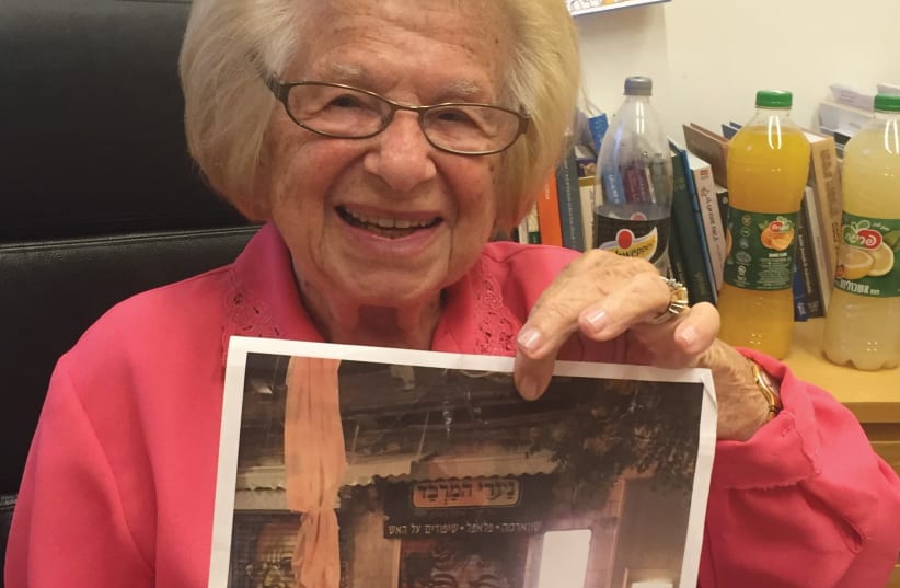 An icon in spraypaint: Dr. Ruth holds up a photo of a storefront in Mahaneh Yehuda that has been decorated with her smiling face (one of the many done by Solomon Souza of influential/historical Jewish figures). (photo credit: ARIEL DOMINIQUE HENDELMAN)