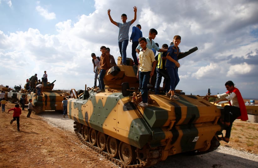 Boys stand on top of a Turkish army vehicle as a military convoy pass by their village on the Turkish-Syrian border line in Reyhanli, Hatay province, Turkey, October 11, 2017.  (photo credit: REUTERS/OSMAN ORSAL)