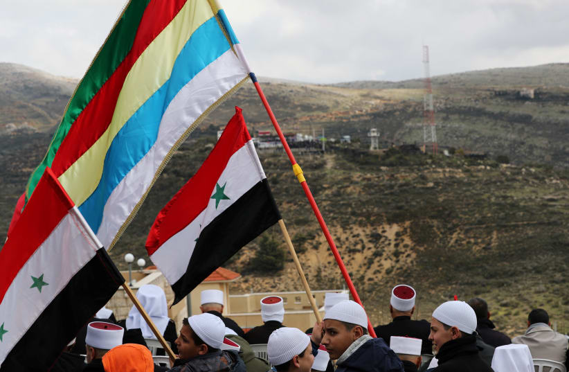 Members of the Druze community holds Syrian and Druze flags as they sit facing Syria, during a rally marking the anniversary of Israel's annexation of the Golan Heights in the Druze village of Majdal Shams. February 14, 2019.  (photo credit: AMMAR AWAD / REUTERS)