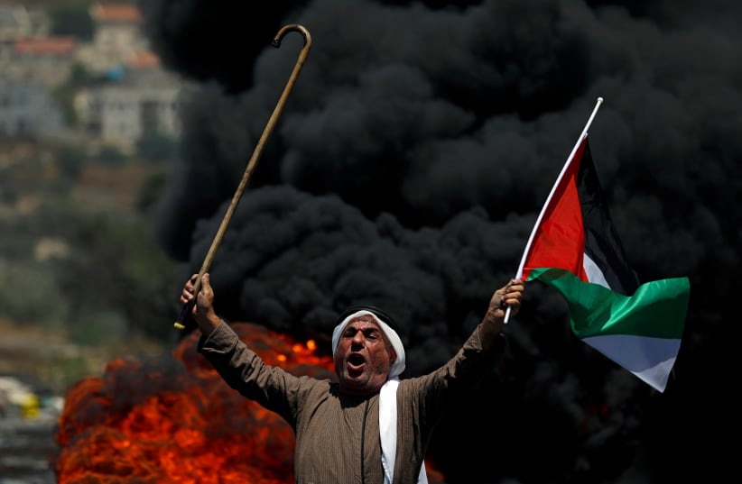 A demonstrator holding a Palestinian flag and a cane reacts in front of burning tires during a protest near the Jewish settlement of Qadomem, in the village of Kofr Qadom in the Israeli-occupied West Bank July 5, 2019 (photo credit: MOHAMAD TOROKMAN/REUTERS)