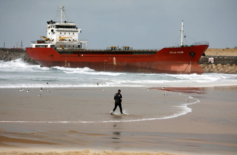 A man walks along the shore of the Mediterranean Sea as a cargo vessel that arrived at Israel's Ashdod port and was swept away when a storm began is seen nearby, close to Ashdod's port in southern Israel December 26, 2019 (photo credit: REUTERS/AMIR COHEN)