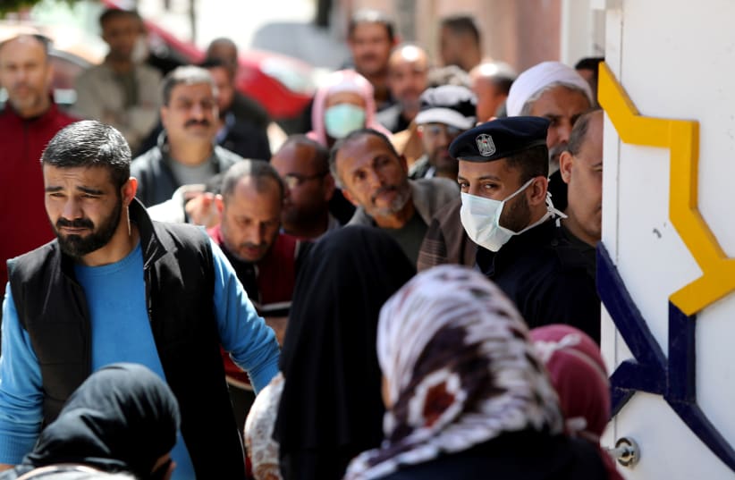 A policeman, wearing a mask as a precaution against the coronavirus disease (COVID-19), stands guard as Palestinians wait outside a bank to withdraw cash, in Gaza City March 29, 2020 (photo credit: REUTERS/MOHAMMED SALEM)