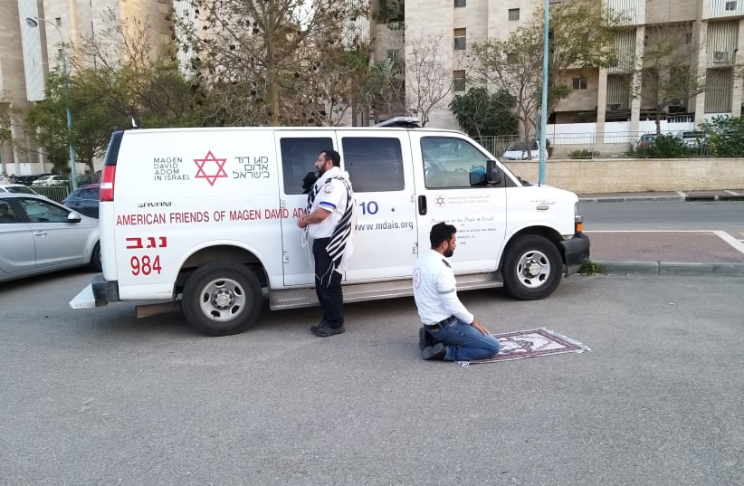 Two paramedics, a Jew wearing a prayer shawl and a Muslim using a prayer mat, pray beside each other near a Magen David Adom (MDA) ambulance in Beersheba, southern Israel, March 24, 2020 (photo credit: MOHAMD ALNBARE/MDA SPOKESPERSON)