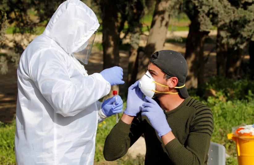 A member of medical staff swaps a Palestinian worker for coronavirus disease (COVID-19) testing, upon his return from Israel, outside the Israeli-controlled Tarqumiya checkpoint near Hebron in the Israeli-occupied West Bank March 26, 2020 (photo credit: MUSSA QAWASMA / REUTERS)
