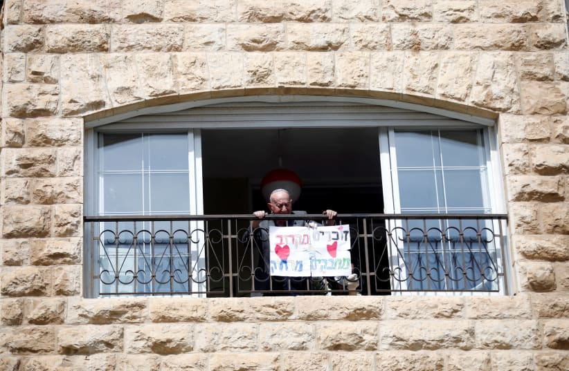 Elias Feinzilberg, a 102-year-old Holocaust survivor, stands at the window of his home in Jerusalem as Israel marks Holocaust Remembrance Day under coronavirus disease (COVID-19) restrictions, April 21, 2020. The Hebrew words on the placard read: "Remembering close-by, embracing from afar." (photo credit: RONEN ZVULUN/REUTERS)