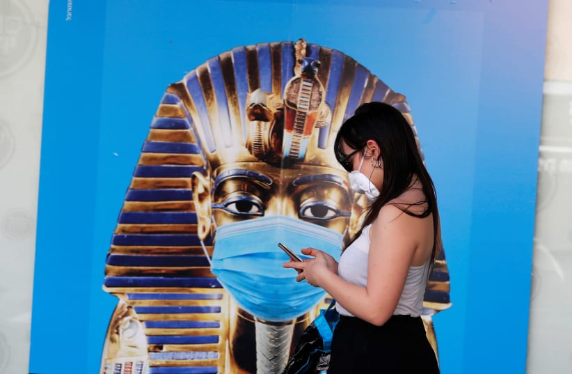 A woman wears a mask and checks her mobile phone as she stands next to a poster in central Tel Aviv, amid the coronavirus disease (COVID-19) restrictions in Israel April 14, 2020 (photo credit: REUTERS/AMMAR AWAD)