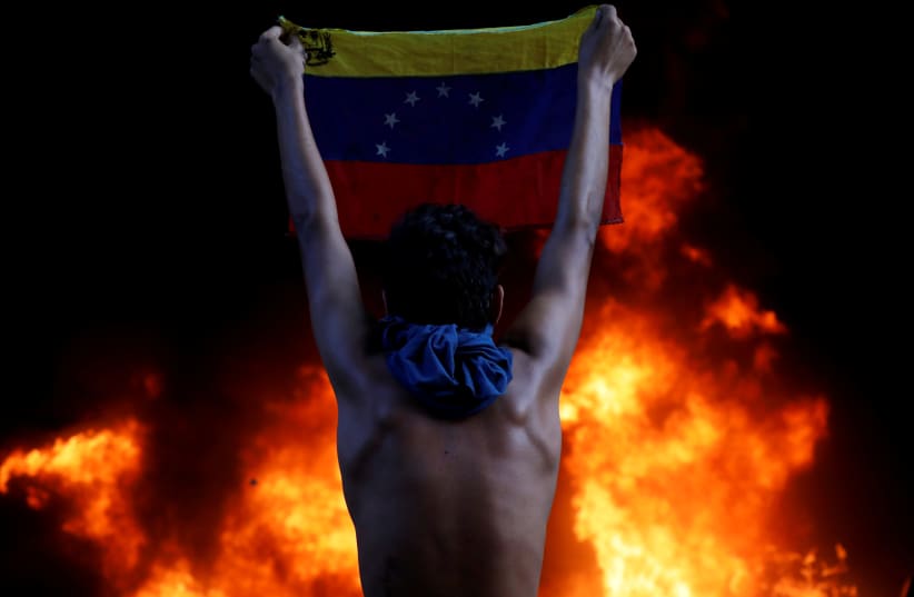 A protestor holds a national flag while standing in front of a fire burning at the entrance of a building housing the magistracy of the Supreme Court of Justice and a bank branch, during a rally against Venezuela's President Nicolas Maduro, in Caracas, Venezuela, June 12, 2017. Protesters angry at t (photo credit: CARLOS GARCIA RAWLINS/ REUTERS)