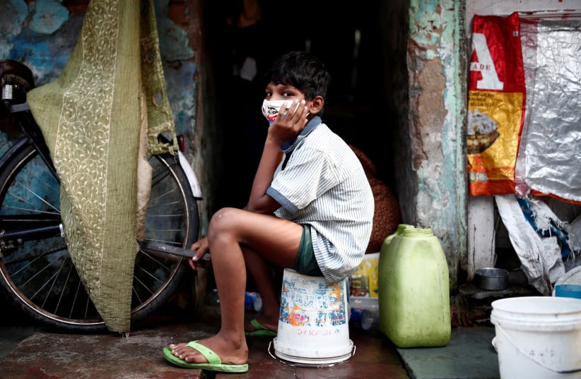 FILE PHOTO: A boy wearing a protective face mask sits on a bucket outside a house in a slum area, during an extended nationwide lockdown to slow the spreading of the coronavirus disease (COVID-19), in New Delhi, India, June 24, 202 (photo credit: REUTERS/ADNAN ABIDI/FILE PHOTO)