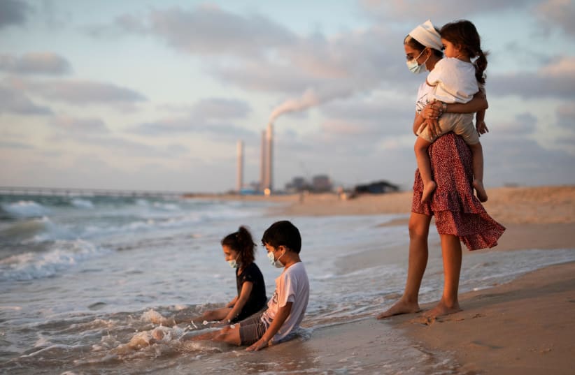 Children wearing masks play in the water along the shore of the Mediterranean Sea as they visit Zikim beach, amid the coronavirus disease (COVID-19) outbreak, in southern Israel July 21, 2020 (photo credit: REUTERS/AMIR COHEN)