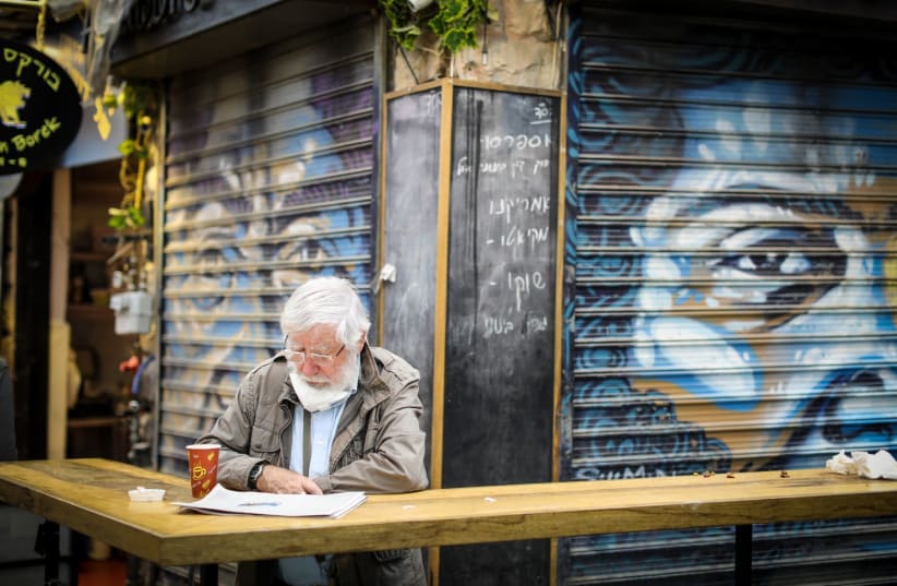 An eldery man is seen sitting alone with coffee and a newspaper at Jerusalem's Mahane Yehuda market amid the coronavirus pandemic, on February 2, 2021. (photo credit: MARC ISRAEL SELLEM/THE JERUSALEM POST)