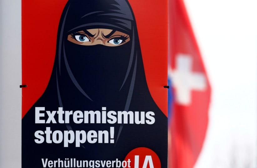 A Swiss national flag flies behind a poster of the initiative committee against wearing the burqa (Verhuellungsverbot) reading "Stop extremism! Veil ban -Yes" in Berikon, Switzerland February 12, 2021. On March 7 Switzerland's voters will decide about a nationwide veil ban. (photo credit: REUTERS/ARND WIEGMANN/FILE PHOTO)