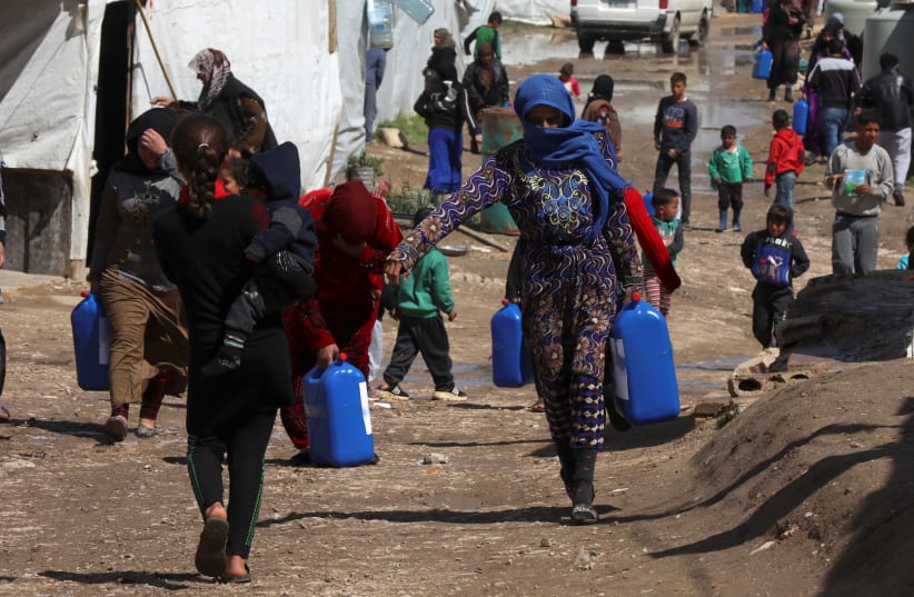 Syrian refugees walk as they carry containers at an informal tented settlement in the Bekaa valley, Lebanon March 12, 2021 (photo credit: MOHAMED AZAKIR/REUTERS)