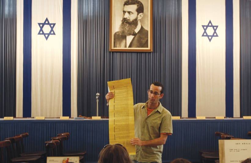 A GUIDE stands under a portrait of Theodor Herzl at Independence Hall in Tel Aviv. Goodman writes about Herzl’s hope that Zionism would save the Jews. (photo credit: GIL COHEN MAGEN/REUTERS)