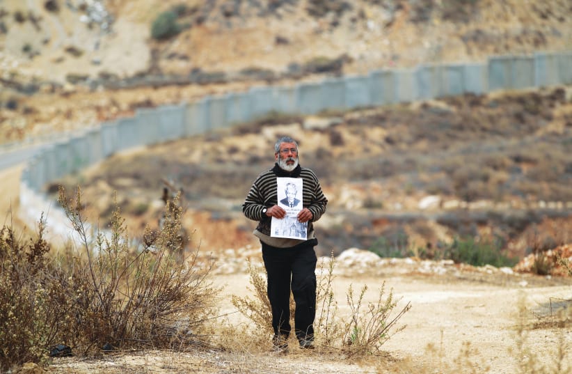 THE SECURITY BARRIER is seen in the background as a Palestinian protester walks with a placard depicting former South African president Nelson Mandela during a demonstration against settlements near the West Bank village of Bil’in in 2013. (photo credit: MOHAMAD TOROKMAN/REUTERS)
