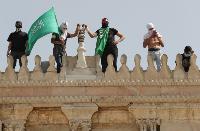 A Palestinian holds a Hamas flag as he stands next to others atop a walk of the al-Aqsa mosque following clashes with Israeli police at the compound that houses al-Aqsa Mosque, known to Muslims as Noble Sanctuary and to Jews as Temple Mount, in Jerusalem's Old City May 10, 2021. (photo credit: AMMAR AWAD/REUTERS)
