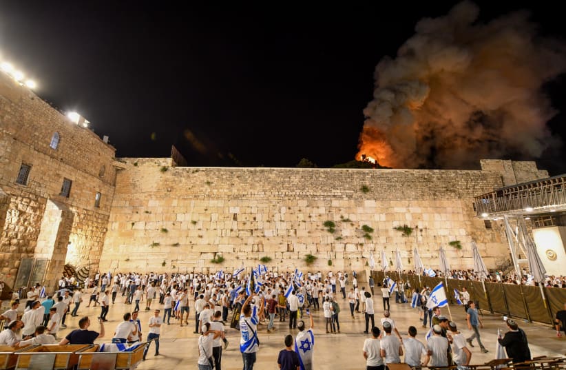 Israelis celebrate Jerusalem day at the Western Wall as blaze is seen in the background at the al-Aqsa mosque compound in Jerusalem Old City, May 10, 2021. (photo credit: MENDY HECHTMAN/FLASH90)