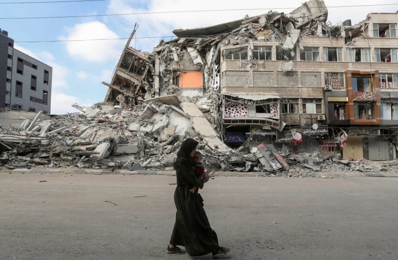 A Palestinian woman carrying her son flees her home during Israeli air and artillery strikes, as she walks near the site of a tower building destroyed in earlier strikes in Gaza City May 14, 2021.  (photo credit: MOHAMMED SALEM/ REUTERS)