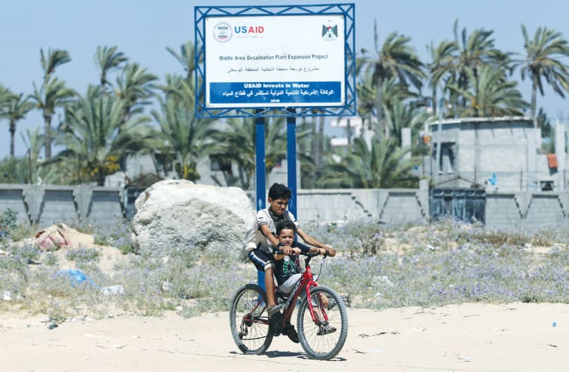 TWO BOYS ride a bike past a USAID sign last week announcing a desalination plant project in the Gaza Strip. (photo credit: IBRAHEEM ABU MUSTAFA/REUTERS)
