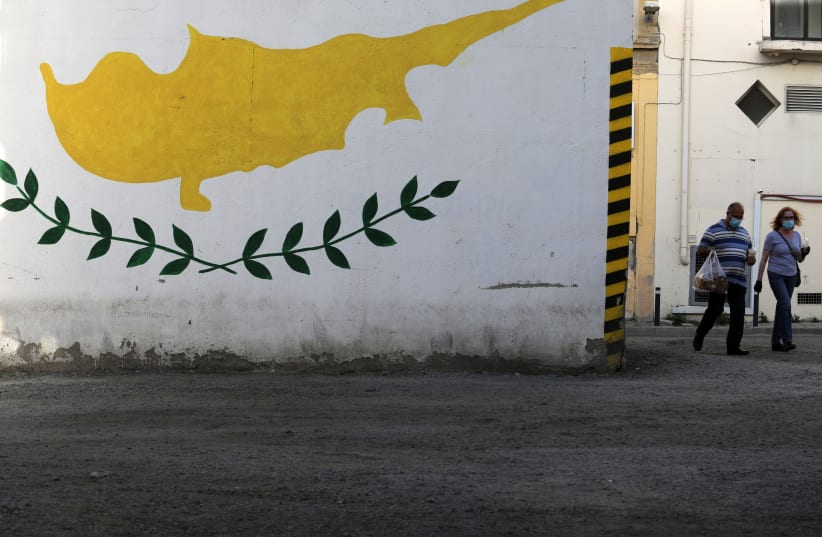  People wearing protective masks walk next to a Cypriot flag painted on a wall in capital Nicosia, Cyprus (photo credit: YIANNIS KOURTOGLOU/REUTERS)