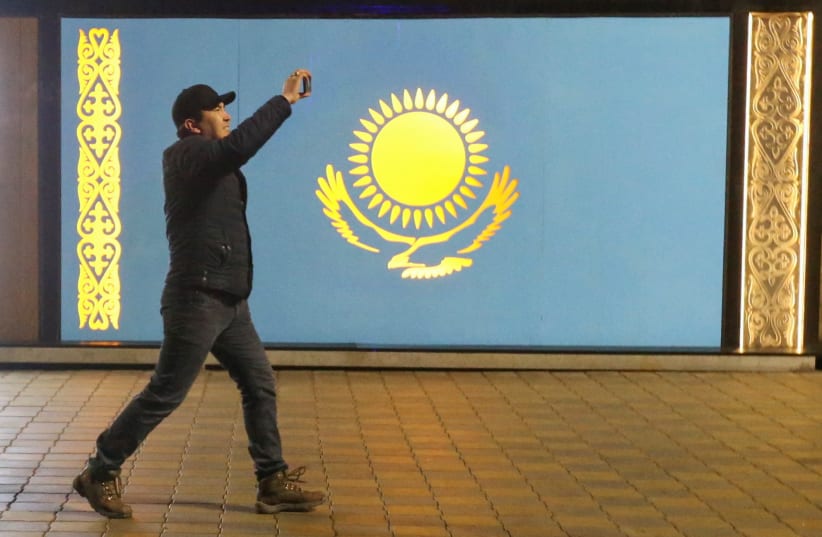  A man holds a mobile phone while walking past a board with a Kazakh state flag during a protest against LPG cost rise following authorities' decision to lift price caps on liquefied petroleum gas in Almaty, Kazakhstan January 5, 2022 (photo credit: REUTERS/PAVEL MIKHEYEV)