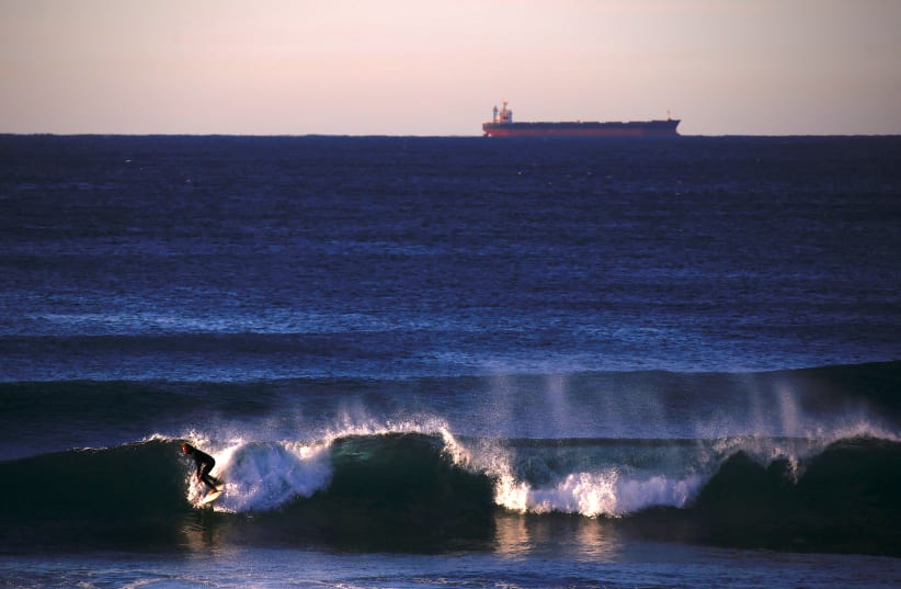  A ship waiting to be filled with a load of coal can be seen behind a surfer riding a wave at Merewether Beach in Newcastle, located north of Sydney in Australia, August 14, 2018 (photo credit: REUTERS/DAVID GRAY)