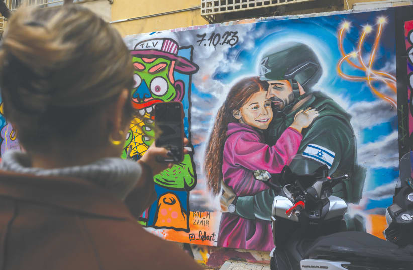  A WOMAN takes a photo of a street art mural of a girl hugging her father dressed in uniform, in Tel Aviv. The national solidarity that was slipping away before the war - and which is so vital to deal with its enormous challenges - has returned. (photo credit: Alexi J. Rosenfeld/Getty Images)