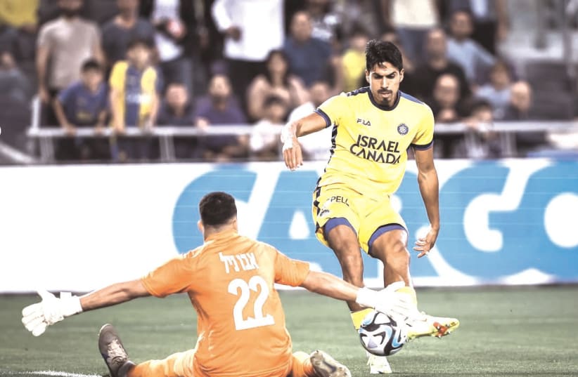 MACCABI TEL AVIV’s Dor Peretz shoots on Bnei Sakhnin ’keeper Mohammed Abu Nil in the yellow-and-blue’s 2-0 win in Israel Premier League action. (photo credit: MACCABI TEL AVIV/COURTESY)