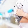 Doctor holds a stethoscope in front of a hospital background (illustrative)