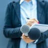 Female reporter holding microphones and writing notes during press conference, media interview, or public relations event.