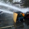 Protesters take cover behind a plywood board as French riot police and gendarmes use a water canon amid clashes during an authorised demonstration organised by labor unions in Paris, October 24, 2026.