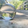 The image shows a clear glass photography sphere (or lensball) placed on a wooden bridge, refracting an inverted view of the trees and sky.