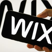 : In this photo illustration, a person holds a smartphone displaying the logo of Wix.com Ltd. (NASDAQ: WIX), a cloud-based web development platform, in front of a screen showing the company's branding on July 31, 2025.