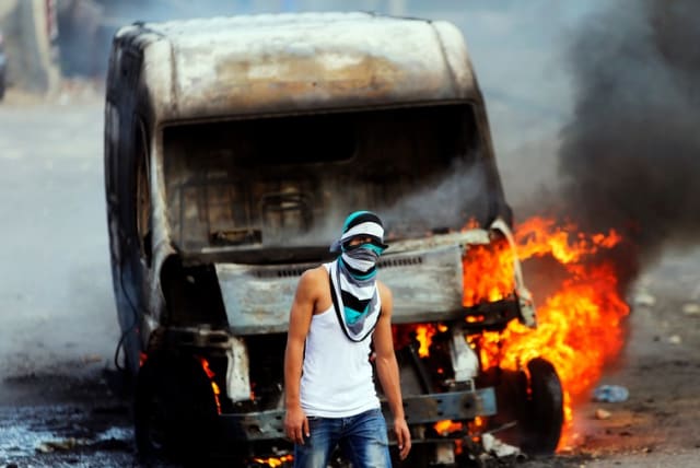 A Palestinian protester stands in front of an Israeli car set torched during clashes with Israeli security forces in east Jerusalem (photo credit: REUTERS)