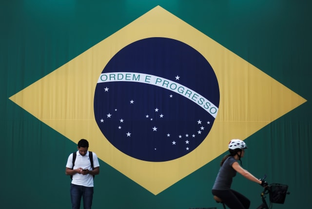 A man checks his mobile phone as a woman riding a bike passes next to a big Brazilian flag in Sao Paulo, Brazil June 28, 2018. (photo credit: REUTERS/NACHO DOCE)