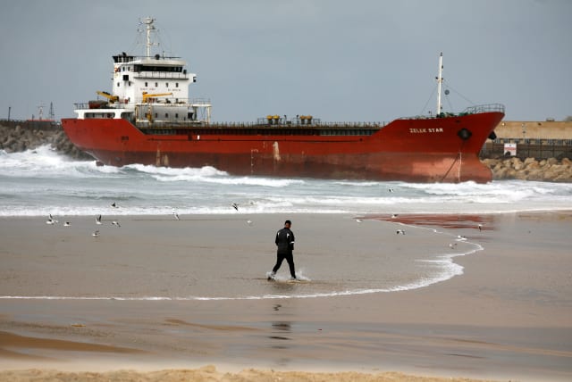 A man walks along the shore of the Mediterranean Sea as a cargo vessel that arrived at Israel's Ashdod port and was swept away when a storm began is seen nearby, close to Ashdod's port in southern Israel December 26, 2019 (photo credit: REUTERS/AMIR COHEN)