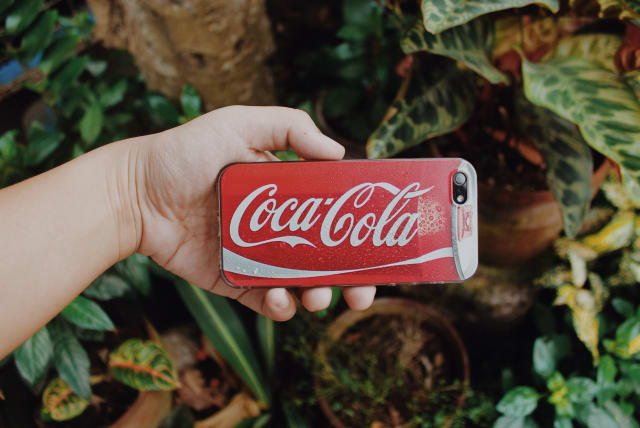  A person holds a phone with a coca-cola branded case. (photo credit: PEXELS)