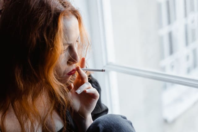A woman smoking while looking out the window. (photo credit: PEXELS)